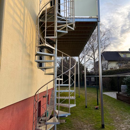 Eine Wendeltreppe ist an der Seite eines Gebäudes befestigt. Sie besteht aus Metall und hat ein Metallgeländer. Das Gebäude hat eine gelb-rote Wand. Die Treppe führt zu einem Balkon mit einem Metallzaun. Es gibt einen Hof mit einem Rasen, einem Baum und einem Haus in der Ferne.