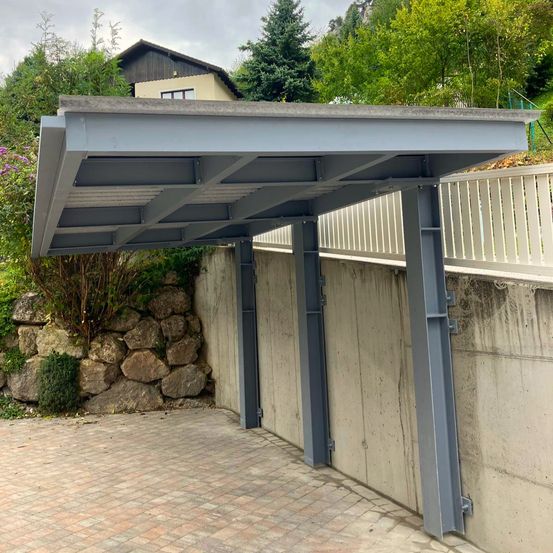 Ein grauer Metallcarport ist an einer Betonwand befestigt und bietet Schatten über einer gepflasterten Einfahrt. Bäume und ein Haus sind im Hintergrund zu sehen.