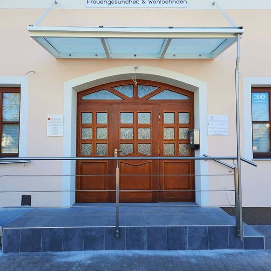 Eine hölzerne Doppeltür mit Glaseinsätzen befindet sich unter einem Glasvordach. Eine Treppe mit Metallgeländern führt zum Eingang. Ein Schild an der Wand lautet Frauengesundheit & Wohlbefinden.