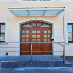 Eine hölzerne Doppeltür mit Glaseinsätzen befindet sich unter einem Glasvordach. Eine Treppe mit Metallgeländern führt zum Eingang. Ein Schild an der Wand lautet Frauengesundheit & Wohlbefinden.