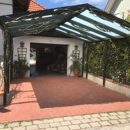 Ein überdachter Carport mit Metallstruktur und durchsichtigem Dach. Es hat einen Ziegelboden, Topfpflanzen und ein rotgedecktes Haus mit offener Garagentür.