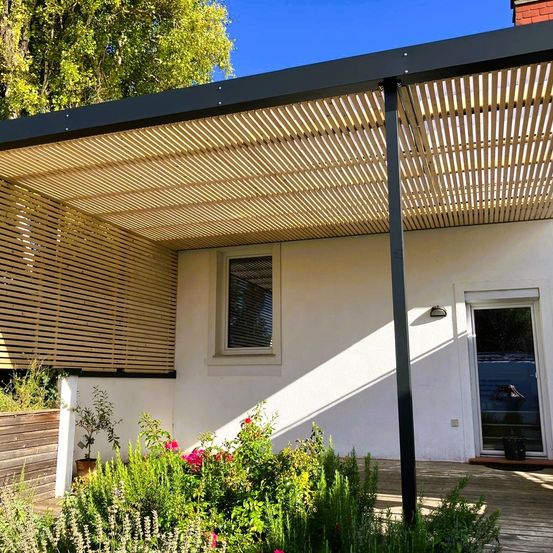 Ein modernes Haus mit Pergola und Glastür. Das Haus hat eine Holzwand und ein Fenster mit Jalousien. Es gibt einen Garten mit Topfpflanzen und Blumen.