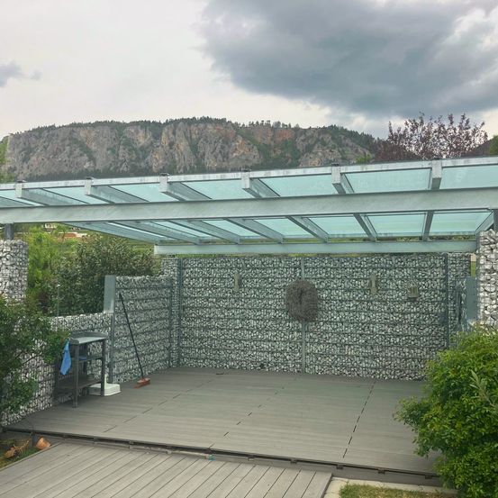 Eine Terrasse mit einer klaren Dachüberdachung und Steinmauern. In der Ferne ein Berg mit viel Grün. Ein Besen und ein weißer Eimer sind auf der Terrasse.
