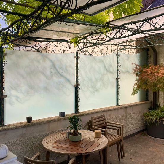 Ein Patio mit einem Holztisch, Stühlen und Pflanzen unter einer Glaswand mit einem Design. Ein weißer Baldachin und ein Baum sind ebenfalls vorhanden.