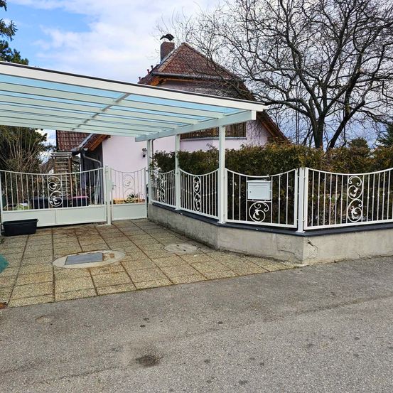 Ein Haus mit einem Ziegeldach und einem überdachten Carport. Der Eingang hat ein weißes Tor und einen Zaun. Bäume und Büsche umgeben das Grundstück.