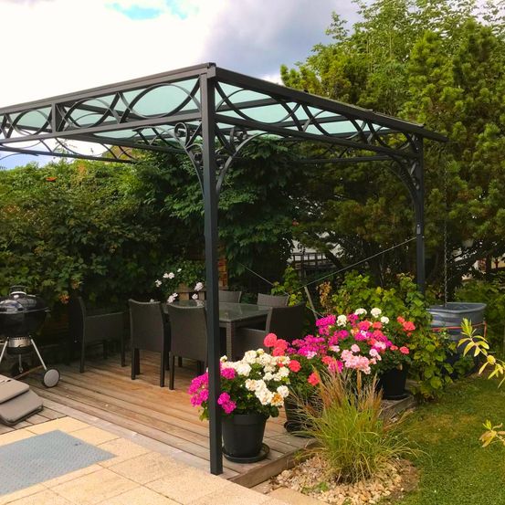 Eine Holzterrasse mit einem schwarzen Metallpavillon und Tisch, umgeben von Pflanzen und Blumen. Ein Grill und Stühle sind links. Bäume und Pflanzen im Hintergrund.