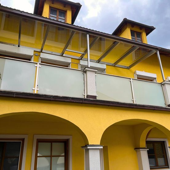 Ein gelbes Gebäude mit einer verglasten Terrasse und Bögen, umgeben von Metallgeländern und Fenstern, unter einem bewölkten Himmel.