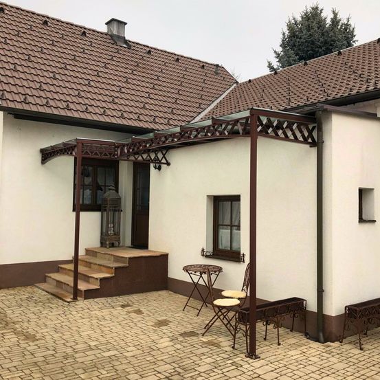 Ein Haus mit einem braunen Dach und weißen Wänden hat einen Innenhof mit Ziegelboden, eine Metallpergola, einen Tisch und Stühle. Das Haus hat ein Fenster, eine Tür und eine Lampe.