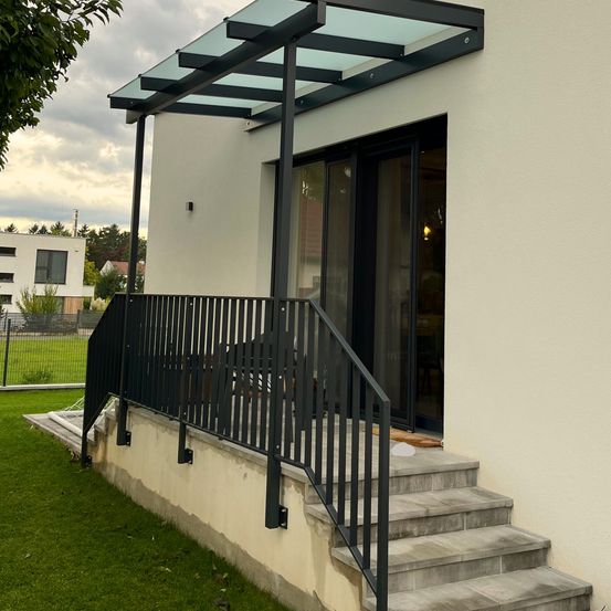 Ein modernes Haus mit einem Glasdach über dem Vorbau, ausgestattet mit schwarzen Geländern und Stufen, die nach oben führen. Der Vorbau hat ein elegantes Design mit Glasfenstern.