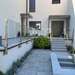 Ein moderner Hauseingang mit einer gepflasterten Einfahrt, Glasgeländern und einer grauen Tür. Pflanzen und ein Topf stehen neben den Stufen.