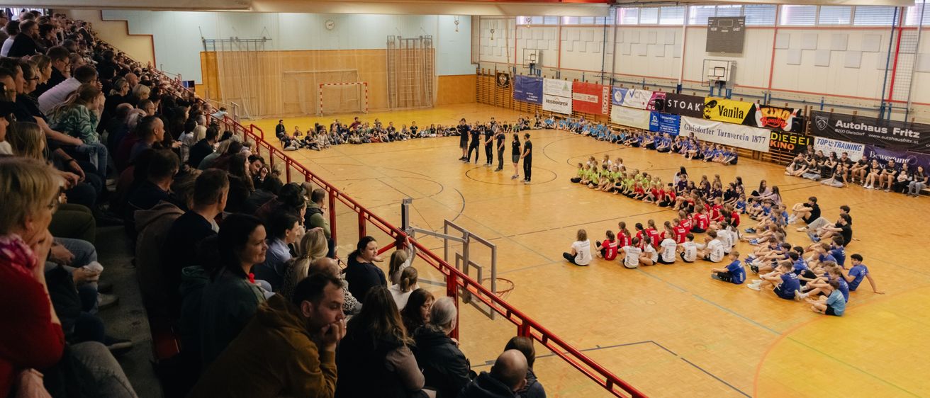 Eine Menschenmenge sieht ein Basketballspiel in einer überdachten Sporthalle. Eine Gruppe von Menschen in grünen Trikots sitzt auf dem Boden.