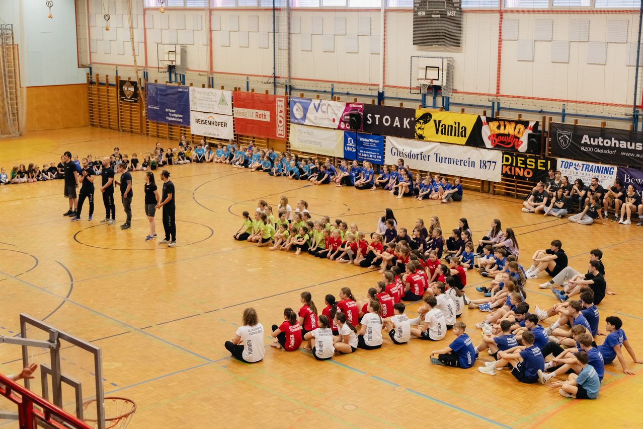 Eine Gruppe von Kindern und Erwachsenen befindet sich in einer Turnhalle und sitzt in einem Kreis um zwei Personen. Sie tragen rote und blaue Hemden mit Namen darauf. An der Wand hängen Banner.