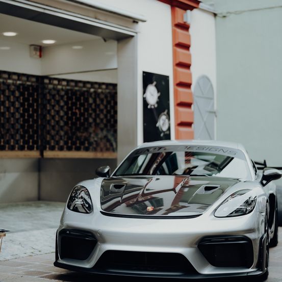 Ein silberner Sportwagen mit schwarzen Akzenten steht vor einem Gebäude mit einer karierten Wand und einer orangefarbenen Säule.