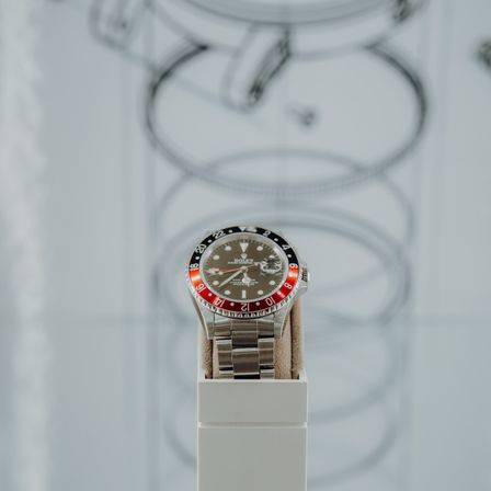 Eine Rolex Uhr mit rotem Zifferblatt und silbernem Ziffernblatt wird auf einer weißen Oberfläche ausgestellt. Das Uhrengesicht zeigt die Zeit als 10:30 Uhr.