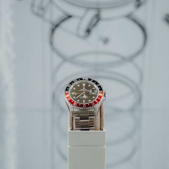 Eine Rolex Uhr mit rotem Zifferblatt und silbernem Ziffernblatt wird auf einer weißen Oberfläche ausgestellt. Das Uhrengesicht zeigt die Zeit als 10:30 Uhr.