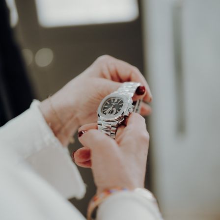 Eine Person justiert eine silberne Uhr mit einem weißen Langarmshirt und Armbändern. Die Uhr hat ein schwarzes Zifferblatt mit weißen Zahlen und einem silbernen Armband.