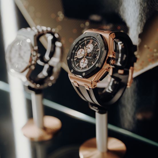 Zwei Luxusuhren werden ausgestellt, eine aus Roségold und Schwarz, die andere aus Silber, beide auf Bronzeständern mit Glasoberflächen.