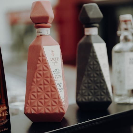 Drei Alkoholflaschen auf einem Regal. Die mittlere Flasche ist terrakottafarben mit einem weißen Etikett mit der Aufschrift Arte Agave Azul. Die anderen beiden Flaschen sind dunkel und durchsichtig.