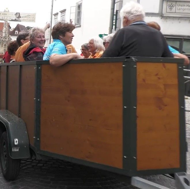 Eine Gruppe von Menschen sitzt auf einem hölzernen Anhänger, der von einem Fahrzeug auf einer Kopfsteinpflasterstraße gezogen wird. Einige lächeln, während andere in die Ferne schauen. Im Hintergrund befinden sich Gebäude und ein Schild.