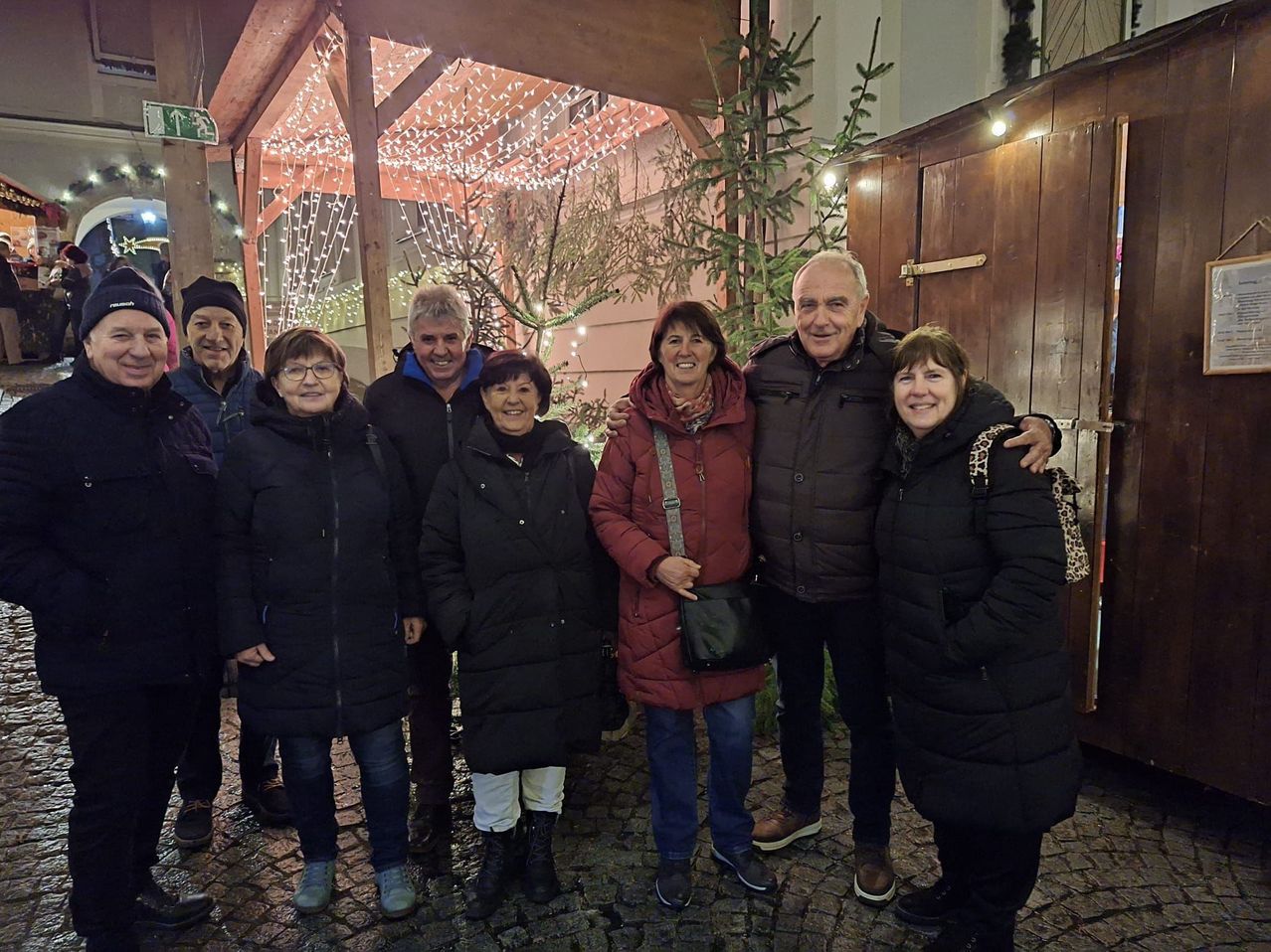 Eine Gruppe Erwachsener in warmen Kleidungsstücken steht vor einer Holzstruktur mit Lichterketten und lächelt für ein Foto.