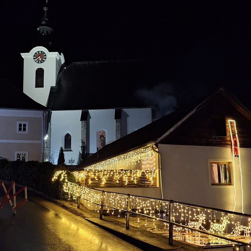 Eine Kirche mit Glockenturm ist nachts beleuchtet. Lichterketten und Weihnachtsdekorationen schmücken das Gebäude. Die Straße ist nass und hat eine Absperrung.