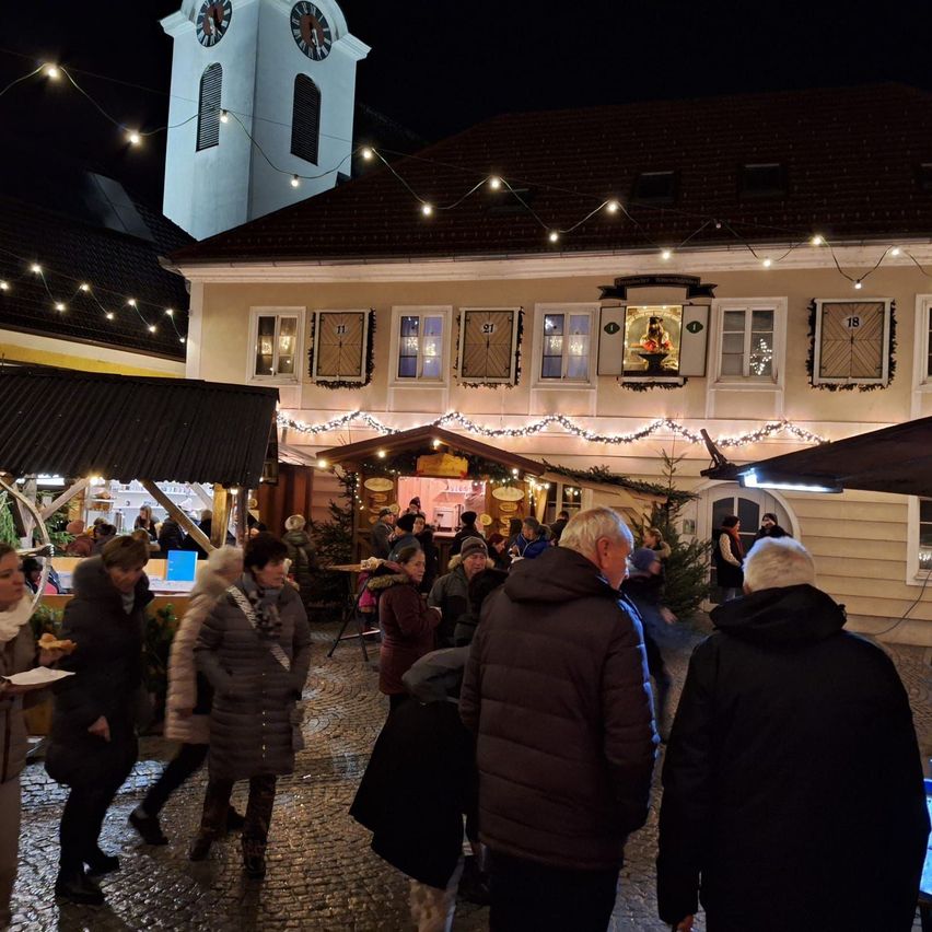 Menschen schlendern bei Nacht durch einen festlich geschmückten Stadtplatz, geschmückt mit Weihnachtslichtern und einem Glockenturm im Hintergrund. Mehrere Menschen versammeln sich bei einem Stand, während andere an Tischen sitzen.