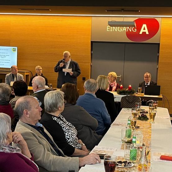 Ein Mann spricht an einem Podium vor einem sitzenden Publikum. Dahinter zeigt eine Projektionsleinwand Text. Kerzen und Getränke stehen auf dem Tisch. Der Raum hat Holzwände und ein Schild, das 'Eingang A' besagt.