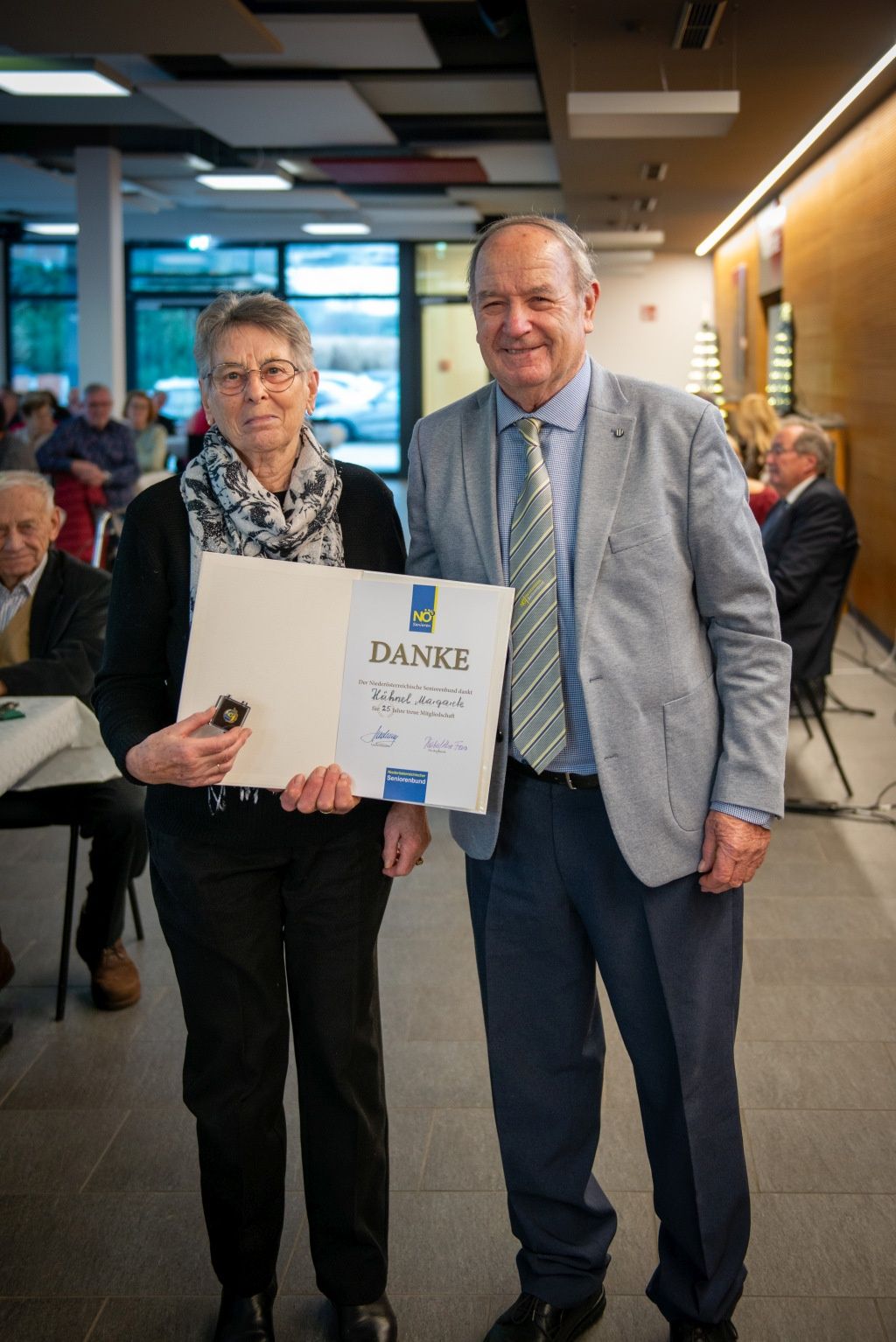 Eine ältere Frau mit Brille und Schal hält eine Urkunde in der Hand, neben einem älteren Mann in Anzug und Krawatte. Sie lächeln beide, und es gibt Menschen, die an Tischen sitzen und im Hintergrund stehen.