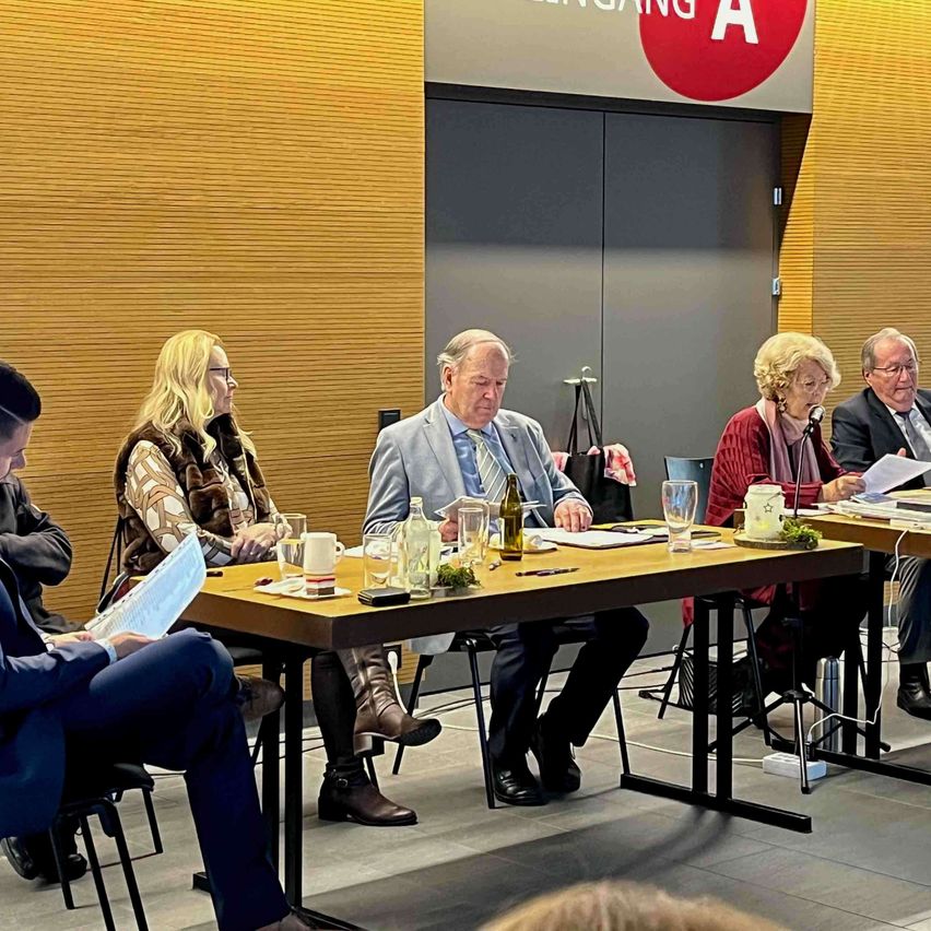 Fünf Personen sitzen um einen Tisch in einem Konferenzraum, jeder mit Papieren und einem Mikrofon vor sich. Ein Schild mit dem Text 'Eingang A' befindet sich über dem Eingang.