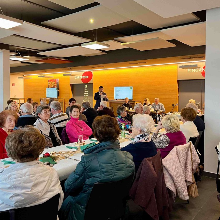 Eine Gruppe älterer Menschen sitzt um einen langen Tisch in einem Konferenzraum. Ein Mann steht an einem Podium mit Mikrofon.