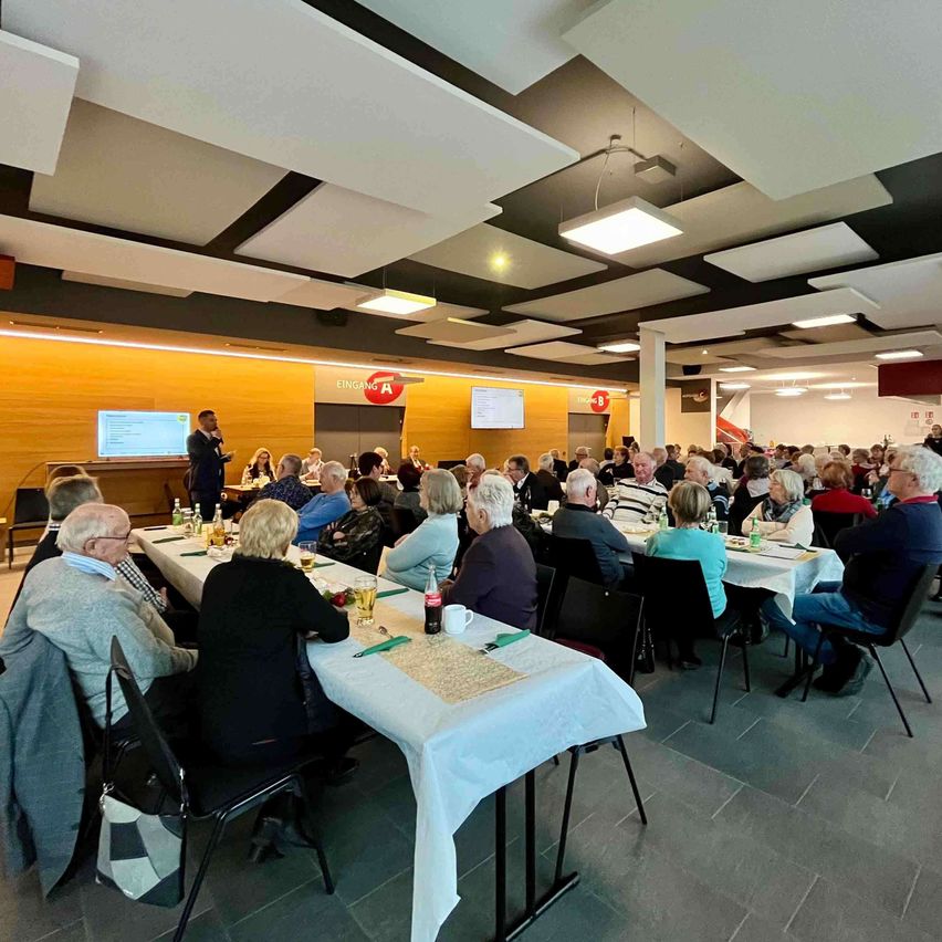 Eine Gruppe von Menschen sitzt um Tische in einem Saal. Eine Person steht an einem Podium. Mehrere Bildschirme und Schilder sind im Hintergrund sichtbar.