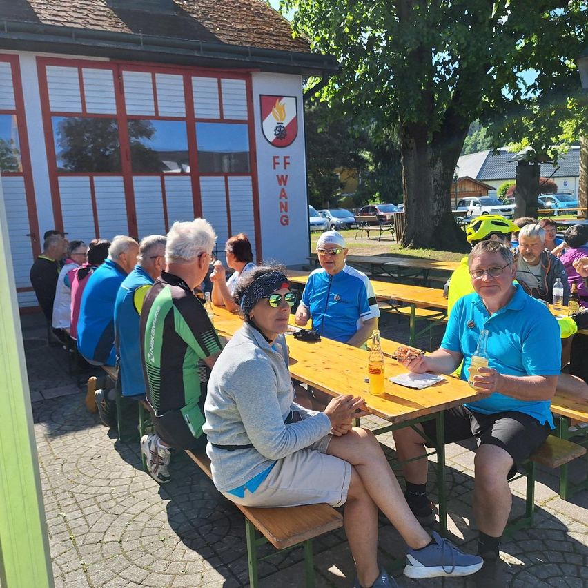 Eine Gruppe von Radfahrern entspannt sich an einer Sitzgelegenheit im Freien vor einem Gebäude, einige mit Sonnenbrille. Flaschen und Essen sind auf dem Tisch.