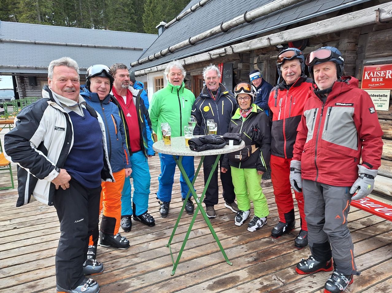 Eine Gruppe von Skifahrern, alle in farbenfroher Winterkleidung gekleidet, versammelt sich um einen kleinen Tisch mit Getränken. Sie stehen auf einer Holzterrasse vor einer Holzhütte mit verschneiten Bergen im Hintergrund.