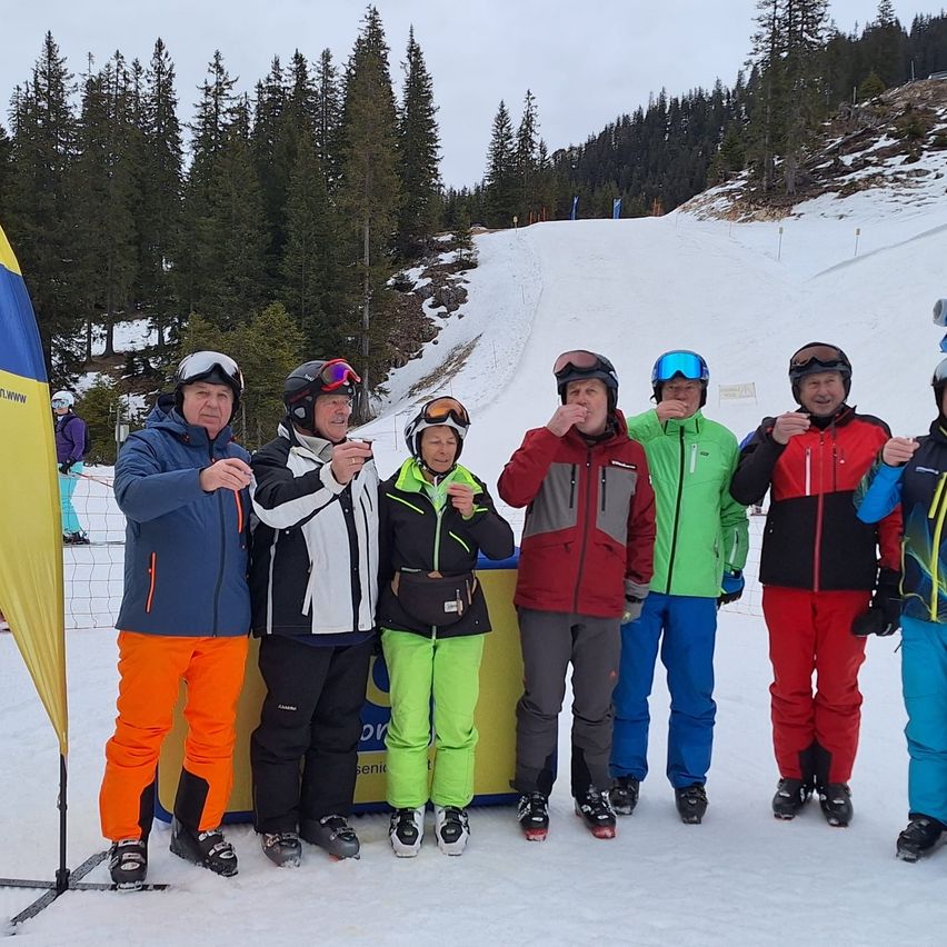 Eine Gruppe von Menschen in Winterkleidung posiert für ein Foto im Schnee, mit Bäumen und einem schneebedeckten Hügel im Hintergrund.