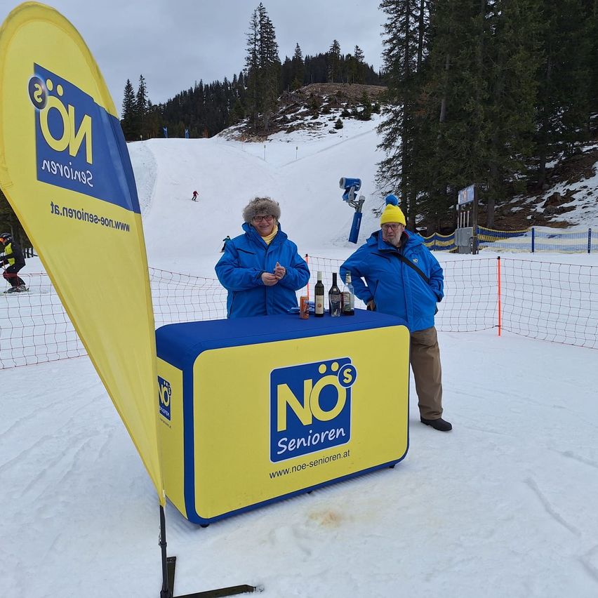 Zwei Personen stehen hinter einem blau-gelben Tisch, wahrscheinlich in einem Skigebiet, tragen blaue Jacken und gelbe Mützen. Ein Skifahrer ist im Hintergrund zu sehen.