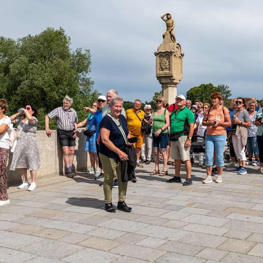 Eine Gruppe von Menschen, hauptsächlich ältere Personen, steht in einem Kreis um eine Statue, einige machen Fotos. Die Statue steht auf einem Podest mit einer Skulptur eines Mannes. Im Hintergrund sind Bäume und ein bewölkter Himmel zu sehen.