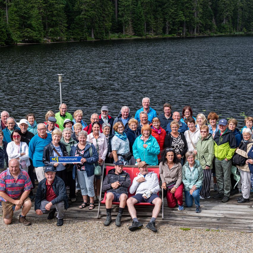 Eine große Gruppe von Menschen, meist älterer Generation, versammelt sich auf einem hölzernen Steg am See, lächelt und posiert für ein Foto. Einige sitzen auf roten Stühlen, und ein Schild mit der Aufschrift 'Blindenmarkt' wird von einer Person in der Mitte gehalten.
