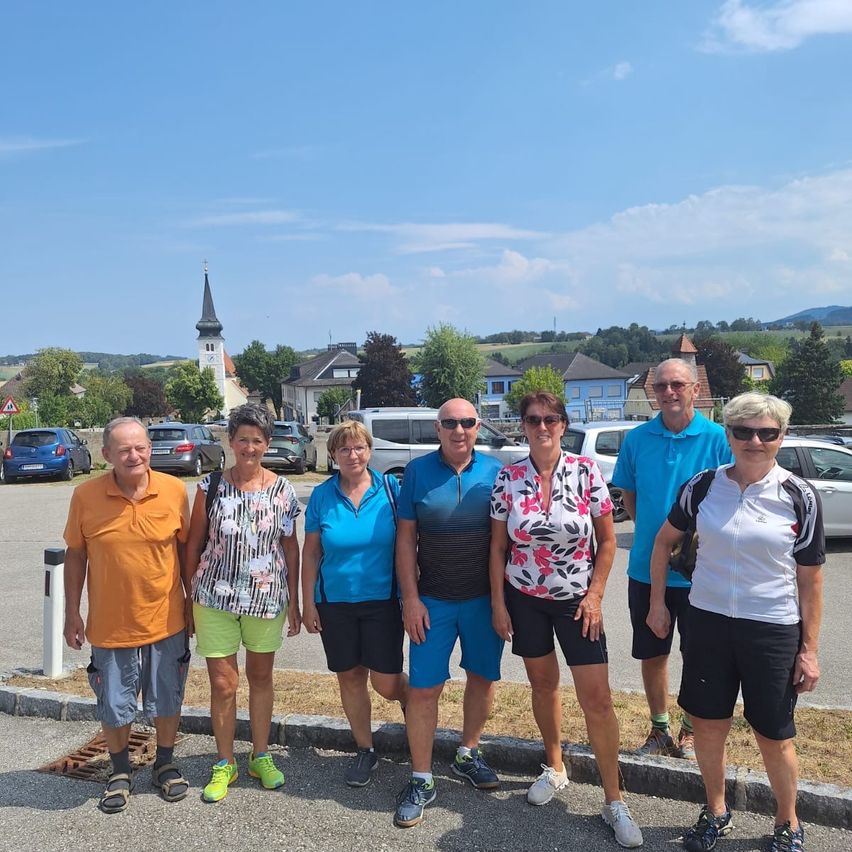 Eine Gruppe von sechs Personen, in Freizeitkleidung gekleidet, steht auf einem Parkplatz mit einem malerischen Blick auf ein Dorf und Berge im Hintergrund.