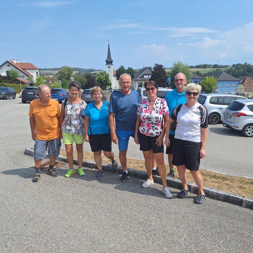 Eine Gruppe von sechs Personen in Freizeitkleidung steht auf einem Parkplatz und lächelt für ein Foto. Hinter ihnen ist ein Kirchturm zu sehen, und mehrere Autos sind geparkt.
