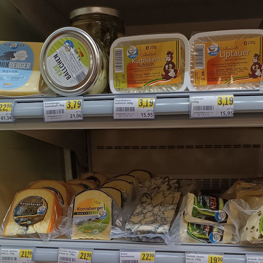 Ein gekühltes Regal zeigt verschiedene Käsesorten, darunter 'Kurbiskern-Außtrisch', 'Liptauer' und 'KONIXBERGER'. Die Preise sind angegeben, wie 3,79€ pro kg für 'Konixberger Käsesäul.180g' und 3,19€ pro kg für 'Kurbiskern-Außtrisch'. Der Käse ist in Plastikverpackungen.
