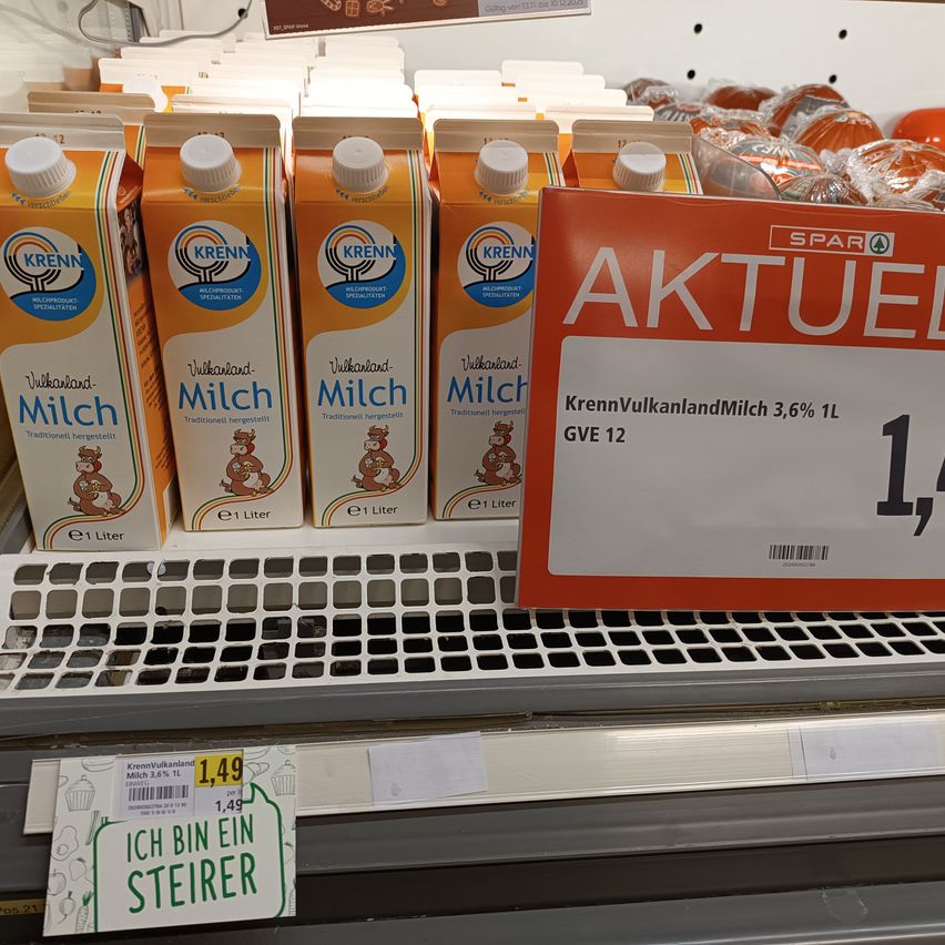 Eine Reihe orangefarbener Milchkartons mit der Aufschrift 'Krenn' wird ausgestellt. Ein Preisschild zeigt 'Aktuell' und einen Preis von 1,49 Euro. Die Milch ist mit 'KrennVulkanlandMilch 3,6% 1L GVE 12' beschriftet.
