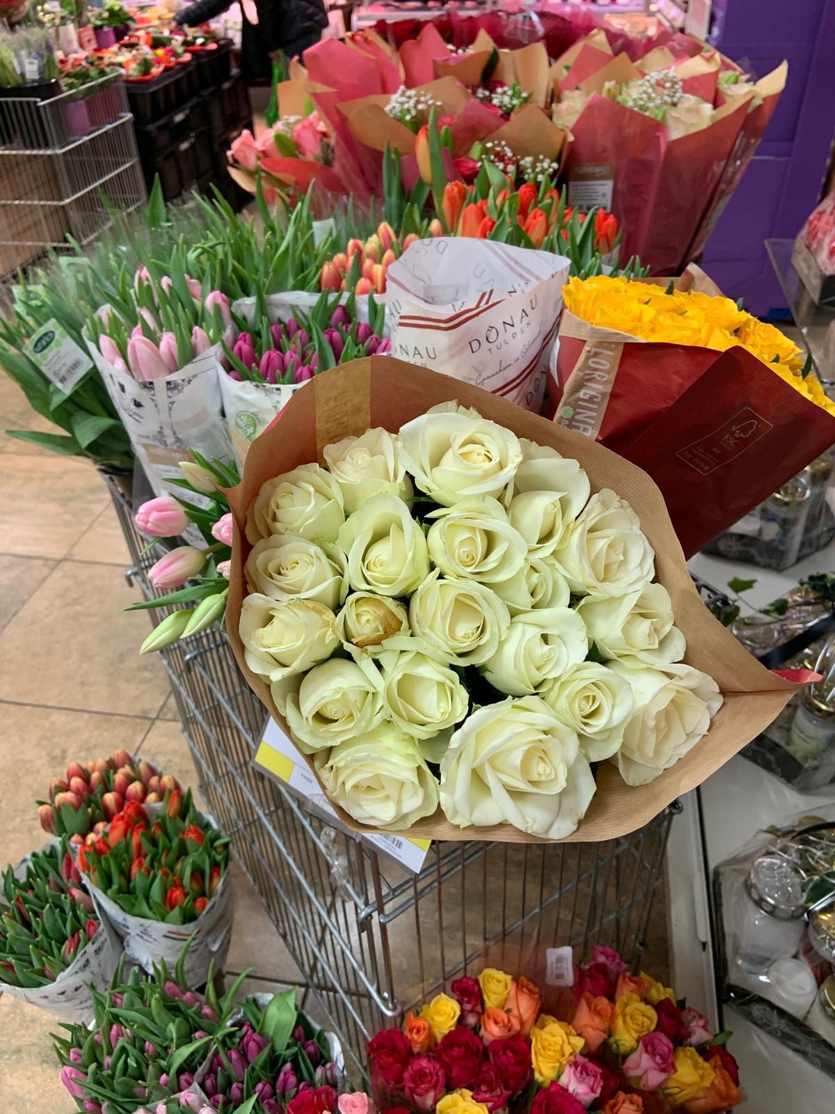 A floral shop displays various flowers. In the foreground, a bouquet of white roses stands out. Surrounding it are tulips in different colors, all neatly arranged.
