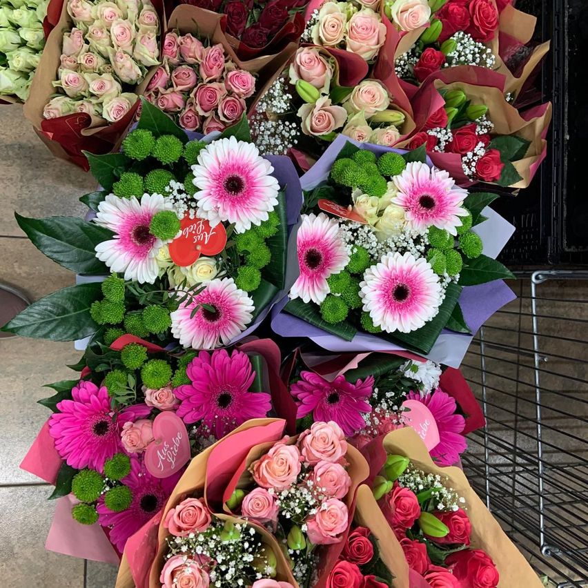 A collection of floral bouquets featuring pink and white flowers, with green leaves and heart tags, arranged on a tiled floor with a metal cart in the background.