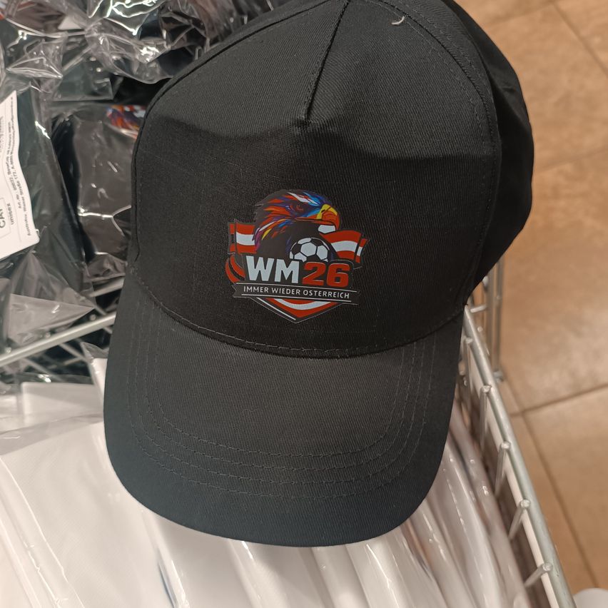 A black baseball cap with WM26 logo is on a rack. The logo features a bird, soccer ball, and flags. It is likely a sports cap.