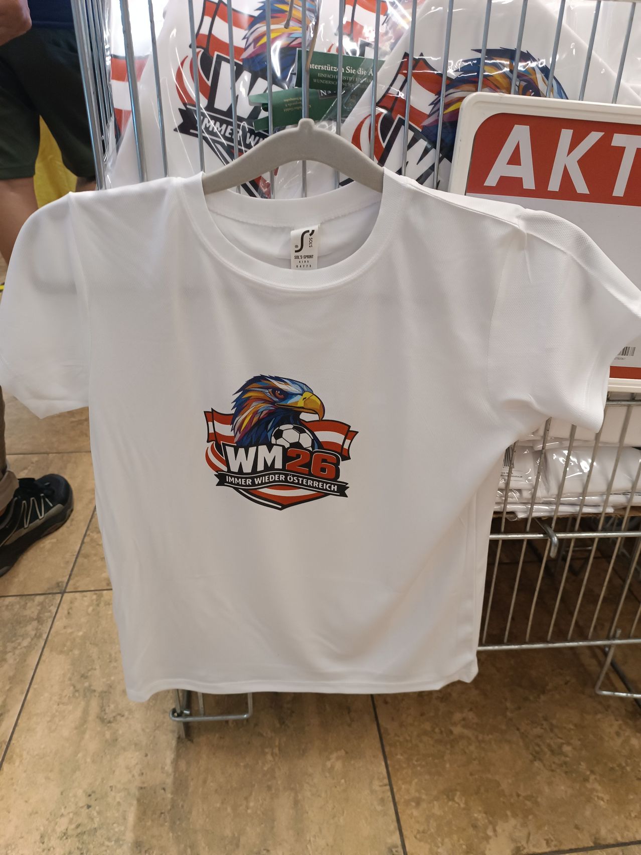 A white shirt with a soccer eagle design is hanging on a hanger in a store. The eagle has a soccer ball and the words 'WM 26' are printed.