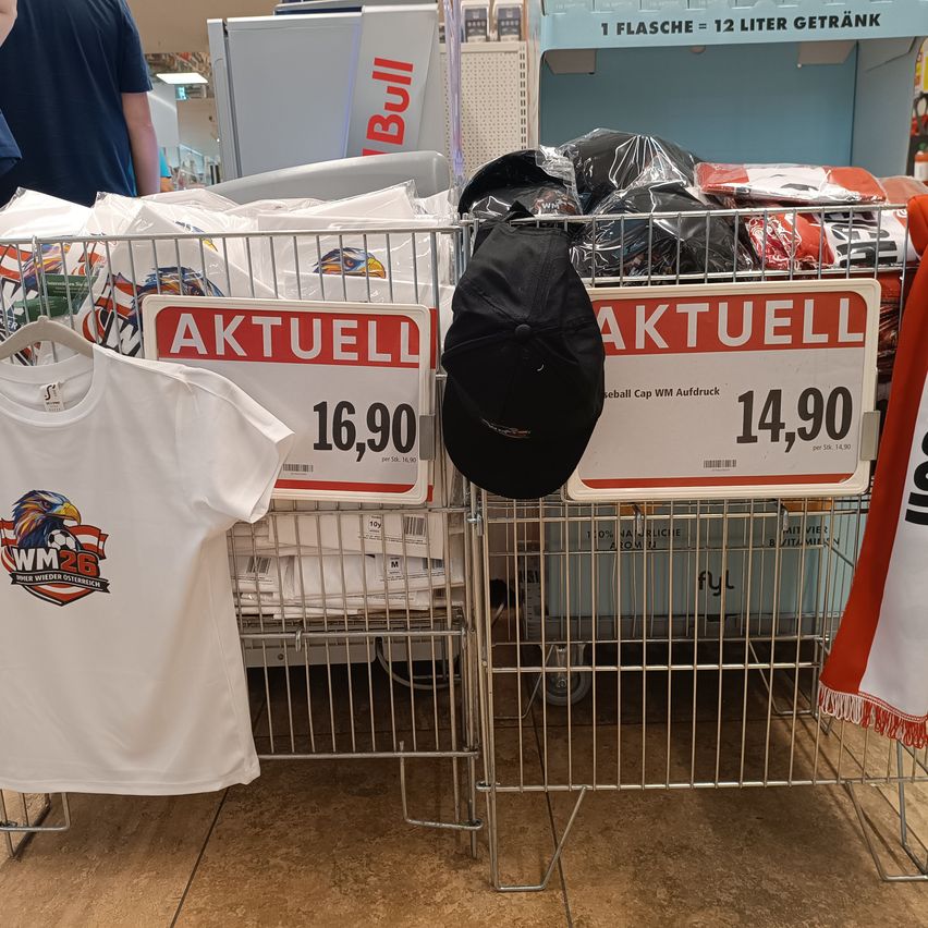 A white shirt with the WM26 logo is on a rack next to a black hat. A shopping cart holds items, including a red sign that says 'Aktuell' and shows the price 16,90.