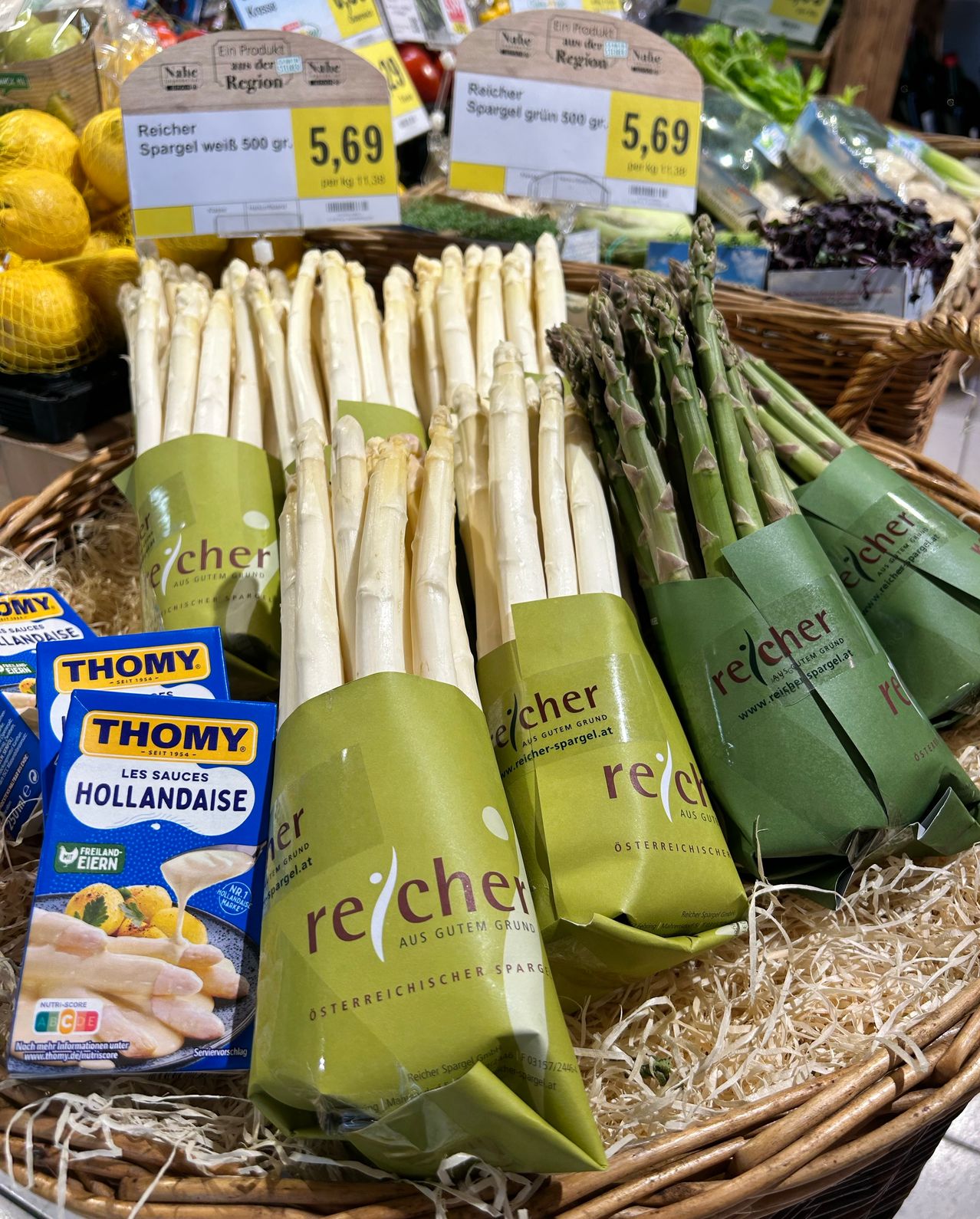 Ein Korb mit Bündeln weißer Spargel, verpackt in grünem re:cher-Papier, zusammen mit Schachteln THOMY Hollandaise-Sauce, auf einem Tisch mit Zitronen und anderem Gemüse.