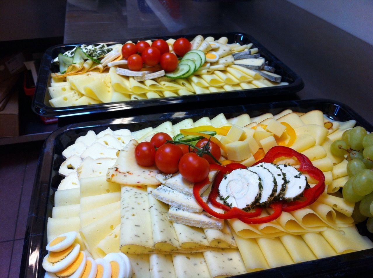 Zwei Tabletts mit verschiedenen Käsesorten, geschnittenen Tomaten, roten Paprika, Gurken und Eiern stehen auf einem Holztisch.