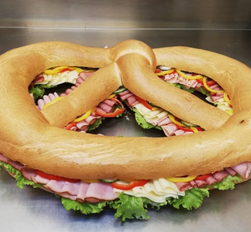 Ein brezelförmiges Sandwich mit Schichten aus Fleisch, Käse und Gemüse wird auf einer Metalloberfläche präsentiert.