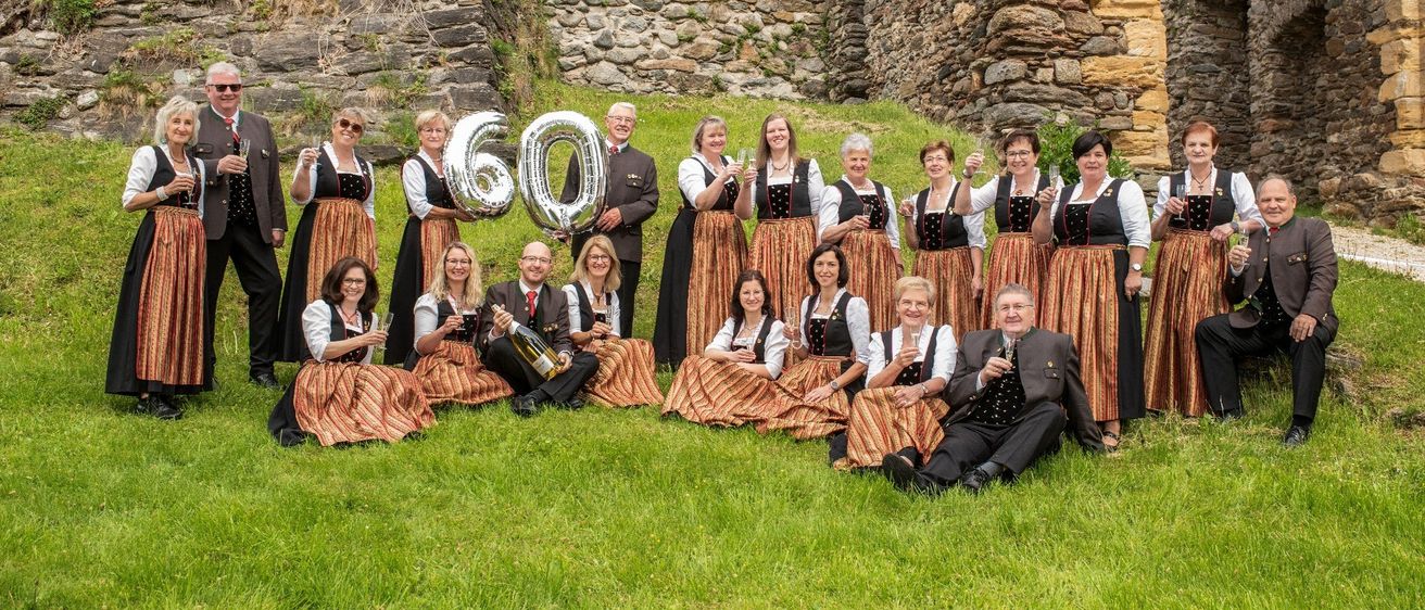 Eine Gruppe von Menschen in traditioneller Kleidung, die vor einer Steinmauer stehen und Weingläser und Luftballons mit der Zahl 60 halten. Ein Mann in formeller Kleidung sitzt auf dem Boden und lächelt.